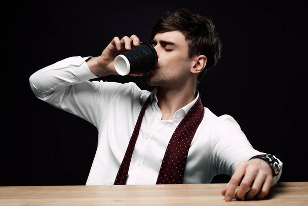 young businessman drinking coffee to go while sitting at wooden desk isolated on black  - Foto, Bild