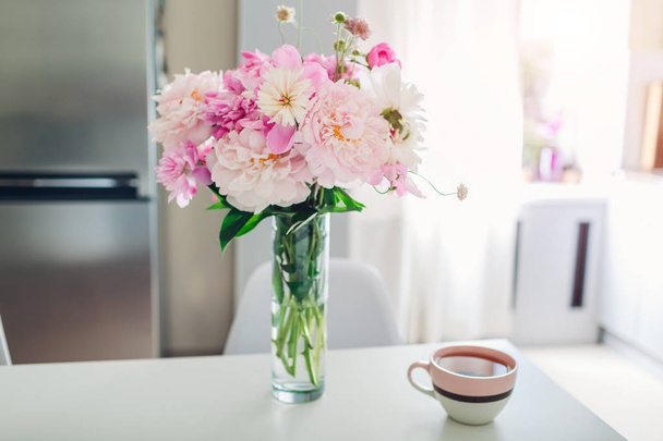 Peonías rosadas con té en la mesa. Diseño moderno de la cocina. Interior de la cocina blanca y plateada decorada con flores
. - Foto, Imagen