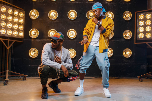 African rappers in caps dance on stage with spotlights on background. Rap performers on scene with lights, underground music, urban style - 写真・画像