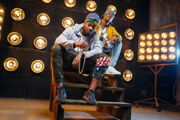Zwei schwarze Rapper sitzen auf den Stufen, performen auf der Bühne mit Scheinwerfern im Hintergrund. Rap-Performer auf der Bühne mit Licht, Underground-Musik, urbanem Stil - Foto, Bild