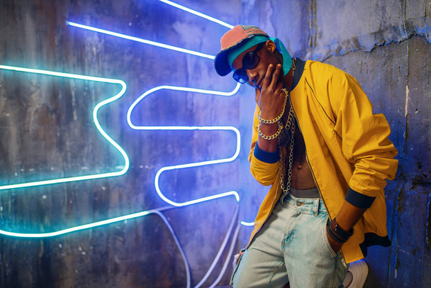 Black rapper in underpass neon lights on background. Rap performer in club with grunge walls, underground - Photo, Image