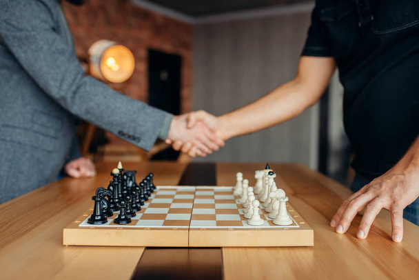 Male chess players shake hands before the game. Two chessplayers begin the intellectual tournament indoors. Chessboard on wooden table - Photo, Image