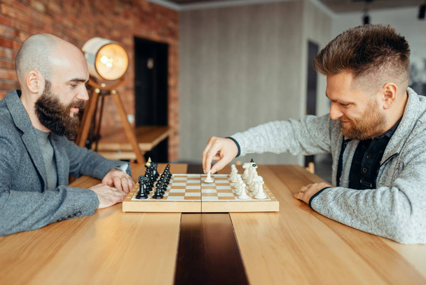 Мужчины шахматисты начинают играть, первый ход. Два шахматиста начинают интеллектуальный турнир в помещении. Шахматная доска на деревянном столе, стратегия игры
 - Фото, изображение