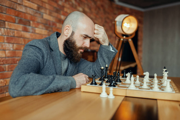 Mannelijke schaakster speelt zwarte figuren, denkproces. Chessplayer aan boord, intellectueel toernooi binnenshuis. Schaakbord op houten tafel, strategie spel - Foto, afbeelding