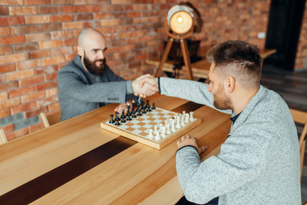 Шахматисты пожимают руки перед игрой. Два шахматиста мужского пола начинают интеллектуальную битву в помещении
. - Фото, изображение