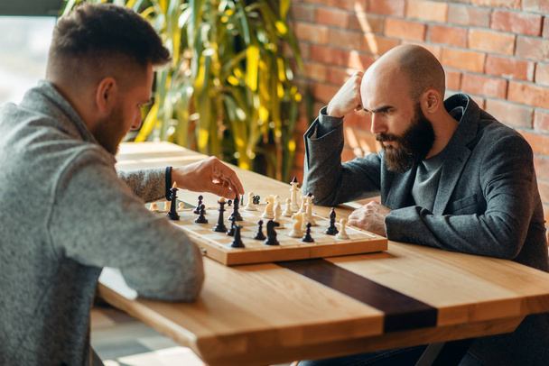 Male chess players playing at board, move the black elephant. Two chessplayers begin the intellectual tournament indoors. Chessboard on wooden table - Zdjęcie, obraz