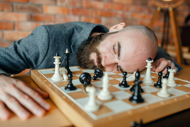 Vermoeide mannelijke schaakspeler slapen op het bord. Chessplayer spelen, intellectuele toernooi binnenshuis. Schaakbord op houten tafel, strategie spel - Foto, afbeelding