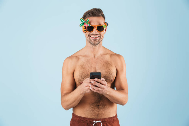 Excited happy adult man in swimwear posing isolated over blue wall background using mobile phone. - Foto, Imagem