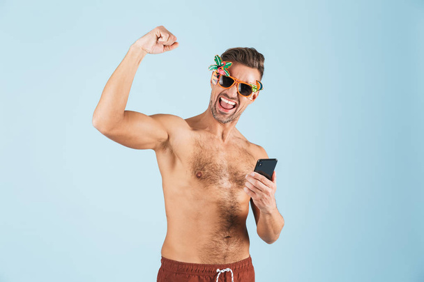 Excited happy adult man in swimwear posing isolated over blue wall background using mobile phone. - Фото, изображение