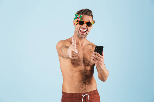 Excited happy adult man in swimwear posing isolated over blue wall background using mobile phone. - Foto, Imagen