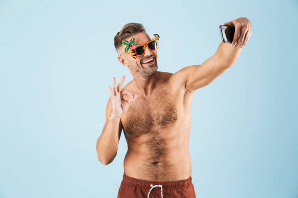 Excited happy adult man in swimwear posing isolated over blue wall background using mobile phone showing okay gesture take a selfie. - Foto, Imagem