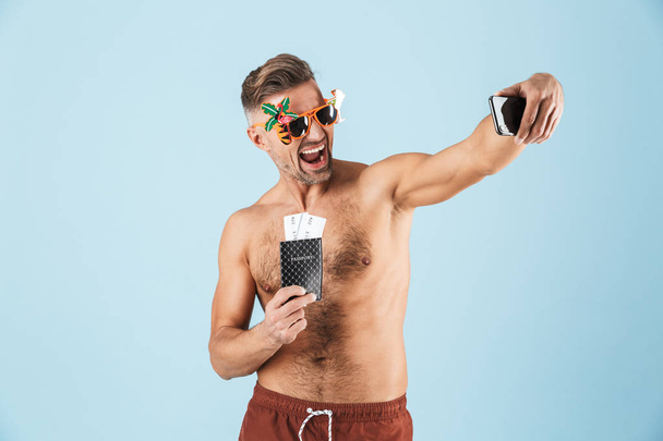 Excited happy adult man in swimwear posing isolated over blue wall background using mobile phone take a selfie. - Fotografie, Obrázek