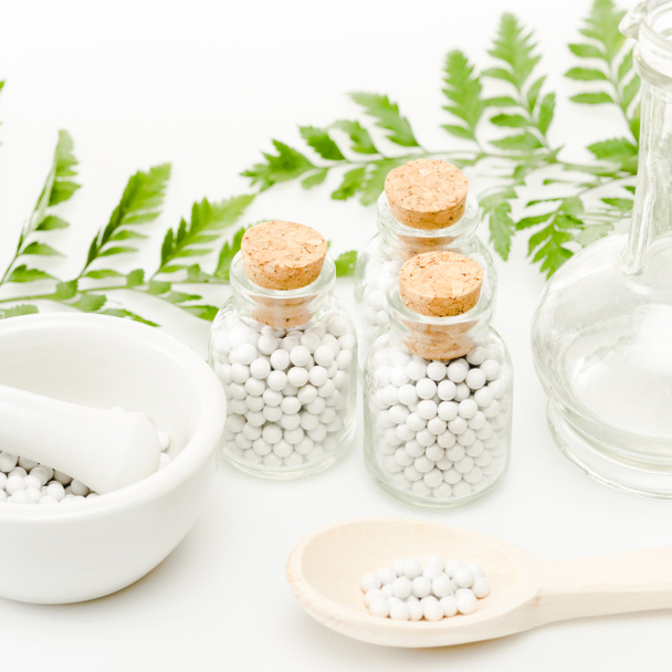 garrafas de vidro com pequenas pílulas redondas perto de argamassa e pilão, colher de madeira, jarra e folhas verdes em branco
  - Foto, Imagem