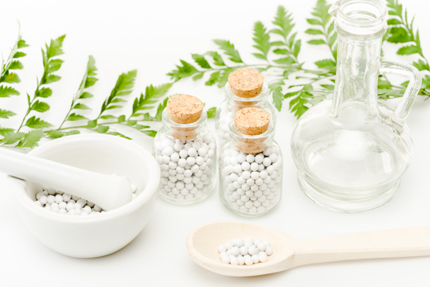 szklane butelki z małymi tabletkami w pobliżu zaprawy i tłuczek, drewniana łyżka, słoik i zielone liście na białym  - Zdjęcie, obraz