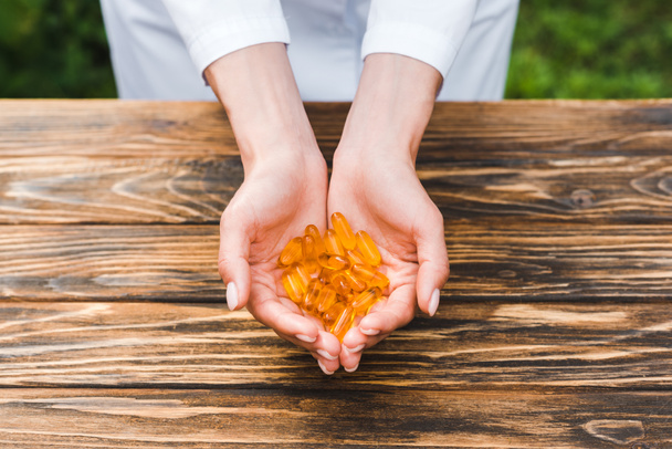 περικομμένη όψη της γυναίκας που κρατά πορτοκαλί χάπια στα χέρια κοντά στο ξύλινο τραπέζι  - Φωτογραφία, εικόνα