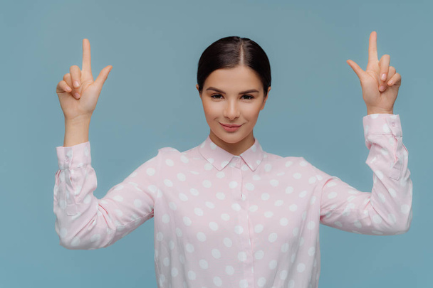 Horizontal shot of self confident brunette young lady in formal shirt points upwards with both fore fingers, shows direction above, dark combed hair, isolated on blue background. Advertisement concept - Photo, Image