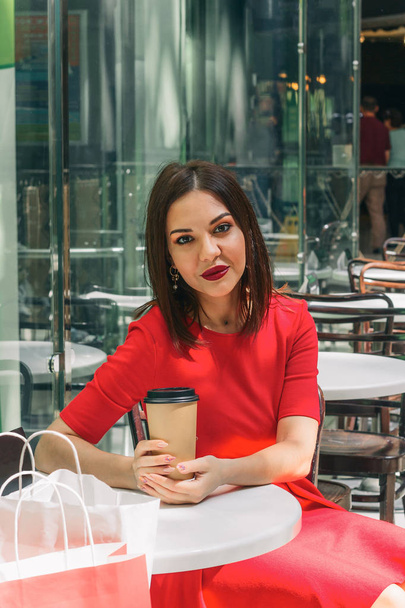 young beautiful brunette woman in red dress drinking coffee in a coffee shop after shopping at the mall - Фото, зображення