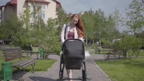 Young happy redhead mother walking with baby carriage and smiling along the street on a nice spring day - Záběry, video