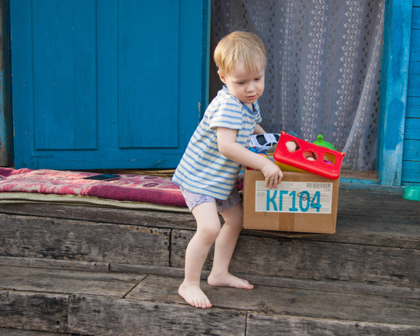 Kora tavasszal, a falu, egy kis szőke fiú, három éves a póló és a bugyi húz egy doboz játékok ki a házból az utcára - Fotó, kép