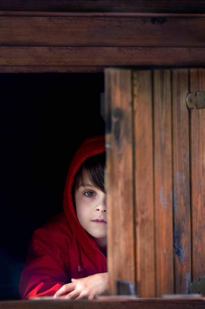 Preteen boy in red sweatshirt, hiding behind a wooden door, look - Foto, Imagem