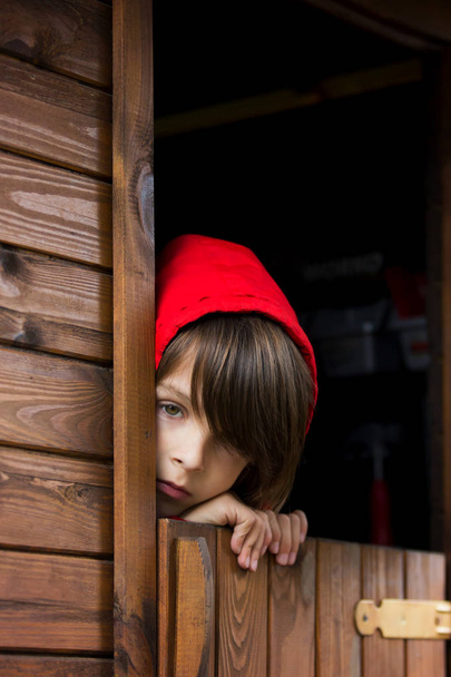 Preteen boy in red sweatshirt, hiding behind a wooden door, look - Φωτογραφία, εικόνα