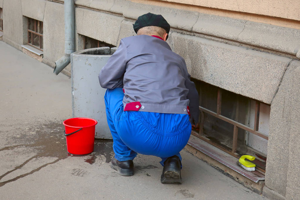 άνθρωπος που κάθεται στην στολή πλένει τα παράθυρα στο υπόγειο. - Φωτογραφία, εικόνα