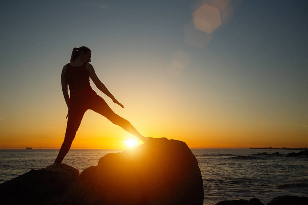 Silhouette of young woman doing fitness exercises on the beach at amazing sunset. Yoga and health. - Photo, Image