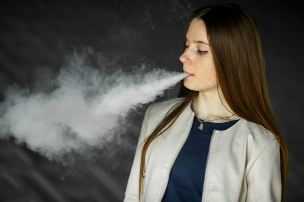 Vape teenager. Young cute girl in  casual clothes smokes an electronic cigarette outdoors in summer day. Bad habit that is harmful to health. Vaping activity. Close up. - 写真・画像