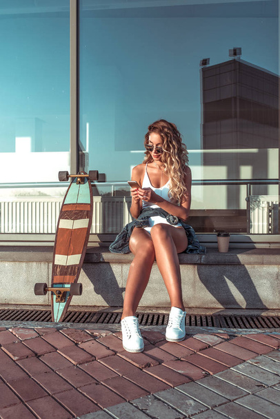 Summer city, woman writes message phone, online internet application, correspondence social networks. Reading news tapes smartphone. Skate longboard, long hair cup coffee tea. Free space for text. - Фото, зображення