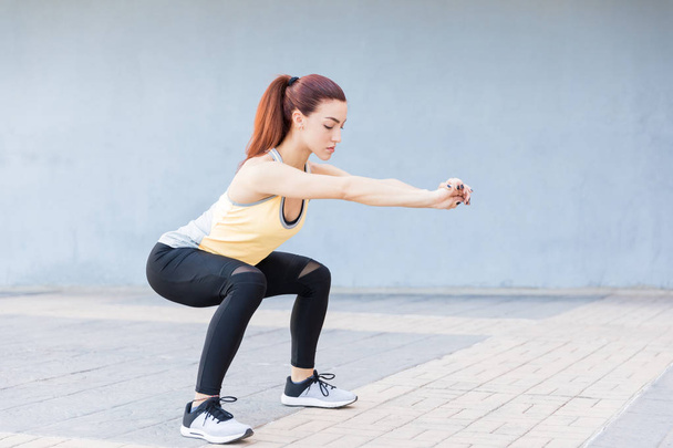 Beautiful woman performing squatting exercise while stretching hands on sidewalk - Zdjęcie, obraz