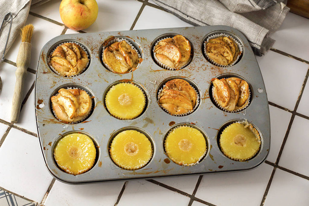 glutenfreie hausgemachte vegetarische Desserts Cupcakes Muffins mit Apfelscheiben und Ananasringen darüber im Backblech in der Küche auf weiß gekachelter Arbeitsplatte, neben einem Küchensieb, Handtuch - Foto, Bild