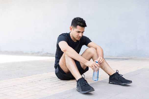 Tired male runner holding water bottle while sitting on footpath in city - Photo, Image