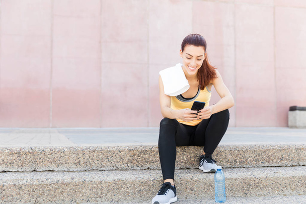 Smiling sporty woman using smartphone after exercise in city - Photo, Image