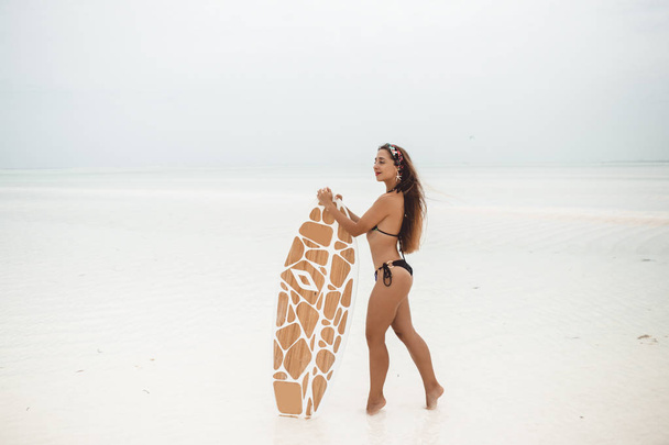 Young sexy woman with sportive tanned body, smiling and jogging with surf board on the beach in cloudly summer day during low tide in Zanzibar - Zdjęcie, obraz