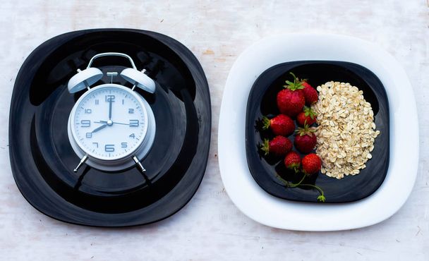 白い背景にオートミールとイチゴの黒いプレート。目覚まし時計は午前8時を示しています。健康的な朝食タイム。健康的な食事の概念. - 写真・画像