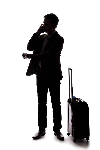 Silhouette of a businessman who is calling for a rideshare with a cellphone.  He is waiting with his luggage as if arriving from an airport.  Isolated on a white background for composites. - Fotoğraf, Görsel