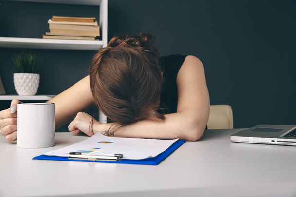 Νυσταγμένη εξαντλημένη γυναίκα στο γραφείο με ένα φλιτζάνι καφέ στο χέρι της. - Φωτογραφία, εικόνα