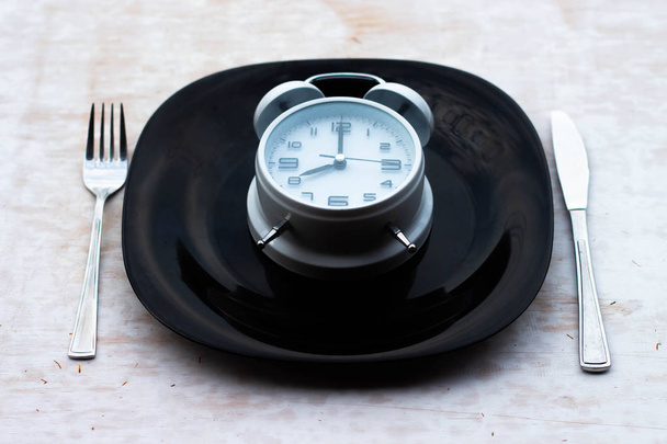 Konzept der Frühstückszeit mit weißem Wecker, schwarzer Platte, Gabel, Messer. das Konzept der gesunden Ernährung. - Foto, Bild