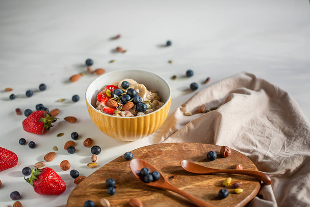 Oatmeal porridge in yellow dish with fruits and nuts on natural wooden tray. Porridge oats with strawberry, pistachios, blueberry, almond and banana. Shadow of sunrise morning. Side view - Photo, image