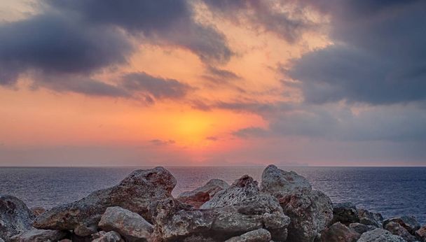 um pôr-do-sol dourado brilhante rodeado por nuvens iluminadas sobre um mar mediterrânico calmo com um primeiro plano de rochas costeiras coloridas pela luz da noite
 - Foto, Imagem