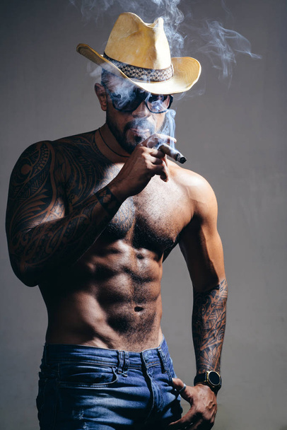 health. smoking cigarette. exhale smoke. bad habit. confidence charisma. sexy abs of tattoo man. male fashion. muscular macho man with athletic body. brutal sportsman in cowboy hat. - 写真・画像