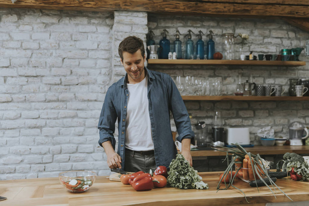 Nuori mies pilkkoo tuoreita vihanneksia maalaismainen keittiö ja valmistaa terveellisen aterian
 - Valokuva, kuva