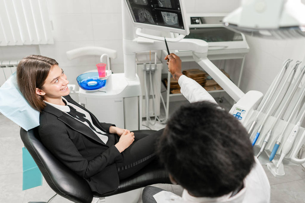 Un joven dentista africano muestra rayos X en el monitor. Mujer en la silla del dentista en la clínica dental. Medicina, salud, concepto estomatológico. dentista tratando a un paciente
. - Foto, Imagen