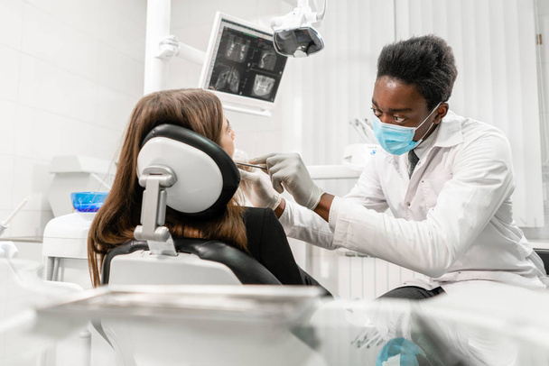 Νεαρός Αφρικανός οδοντίατρος με ασθενή. Γυναίκα στην οδοντιατρική καρέκλα στην οδοντιατρική κλινική. Ιατρική, υγεία, στοματολογία. οδοντίατρος που θεραπεύει ασθενή. - Φωτογραφία, εικόνα