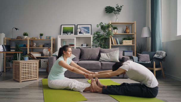 Мужчина и женщина делают упражнения на растяжку, держась за руки, сидя дома на ковриках
 - Кадры, видео