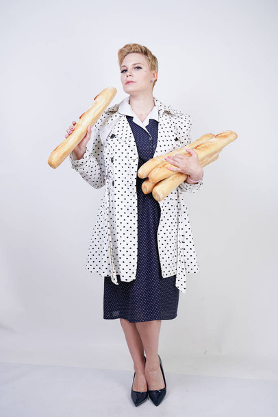 okouzlující růžová žena s krátkými vlasy v jarním kabátě s polky a s bagety a užívajícími si je na bílém pozadí ve studiu. a velikostní dívka v retro šatech s chlebem. - Fotografie, Obrázek