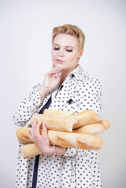 γοητευτικό pinup γυναίκα με κοντά μαλλιά σε ένα παλτό άνοιξη με πουά ποζάρει με μπαγκέτες και απολαμβάνοντας τους σε ένα λευκό φόντο στο Studio. συν μέγεθος κορίτσι σε ρετρό ρούχα κρατώντας ψωμί. - Φωτογραφία, εικόνα
