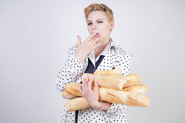 charming pinup woman with short hair in a spring coat with polka dots posing with baguettes and enjoying them on a white background in the Studio. plus size girl in retro clothing holding bread. - Foto, afbeelding