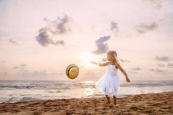 Cute toddler girl with blonde hair in a white tutu dress and hat walking on a sandy beach at sunset. Happy childhood memories, Summer vacations , travel concept - Foto, Bild