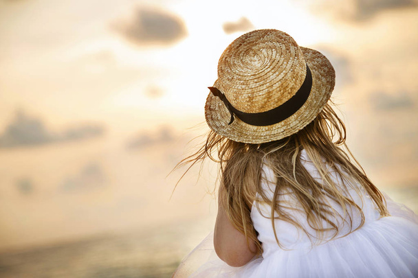 Pretty little girl from behind with long blonde hair in a straw hat and a white tutu dress sitting on a summer beach. Sunny day, sunset, dramatic sky - Foto, immagini